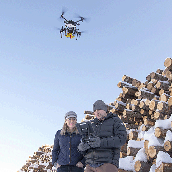 Two people with a drone remote stand in front of a wood pile with a drone in the background